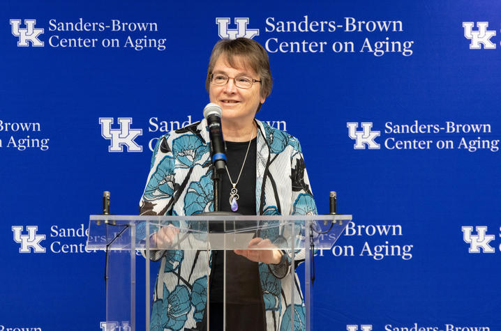 Linda Van Eldik, Ph.D., speaks at a podium in front of a Sanders-Brown Center on Aging backdrop during a University of Kentucky event.