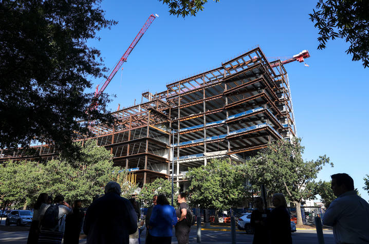 A steel-framed building under construction rises several stories high, with a large red crane extending above it against a clear blue sky. In the foreground, a small group of people stands on the sidewalk beneath trees, looking toward the construction.