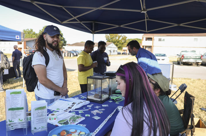 A community outreach event taking place under a blue canopy tent. A table covered with a blue tablecloth displays brochures, stickers, and small containers, while two seated participants interact with a standing attendee wearing a backpack. 
