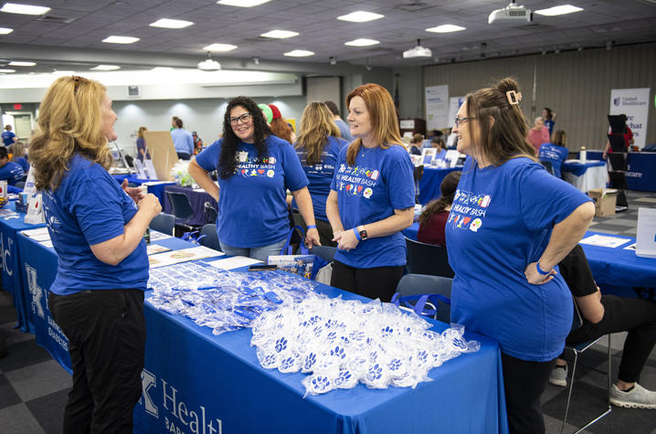 Be Healthy Bash volunteers gather at an information table during the community wellness event. Four people wearing blue Be Healthy Bash shirts stand and talk around a display table inside a large event hall. Tables, exhibits and attendees are visible throughout the room.