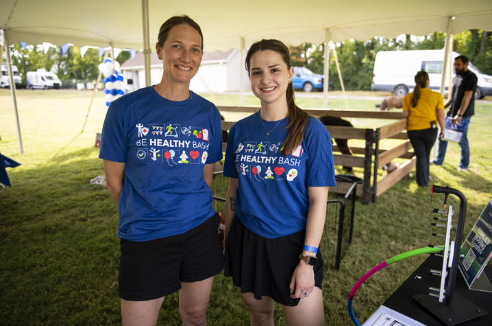 Be Healthy Bash participants stand near an activity area under an event tent. Two people wearing blue Be Healthy Bash shirts pose inside an event tent. A fenced animal exhibit and activity stations are visible in the background.