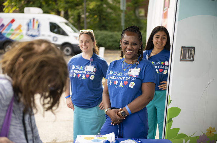 Be Healthy Bash volunteers welcome attendees at a mobile health unit. Three people wearing blue “Be Healthy Bash” shirts stand beside a mobile health unit and greet an attendee at an outdoor event. A table with event materials is in the foreground, and a branded vehicle is visible in the background.