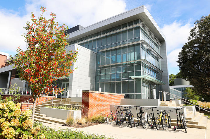 A modern, multi-story glass academic building sits on a landscaped campus, with a small tree showing autumn colors in the foreground and a bike rack filled with bicycles near the entrance on a sunny day.
