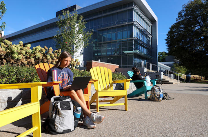 Student sitting in a bright yellow Adirondack chair outside a modern glass campus building, working on a laptop with a backpack and water bottle nearby, while other students sit and study along the walkway on a sunny day.