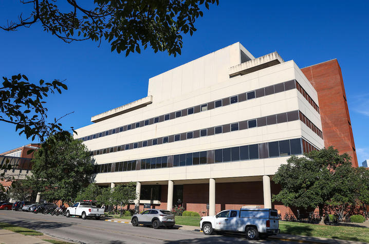 This is an exterior photo of the College of Nursing Building at UK, a multistory building with a white stone front lined with columns along a street with parked vehicles and a clear blue sky. 