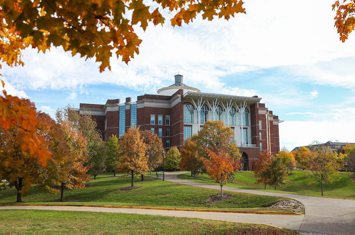 A large brick academic building sits on a gently sloping hill, surrounded by trees with bright autumn foliage in shades of orange, red, and yellow. A winding paved path curves through the grassy landscape under a partly cloudy sky.