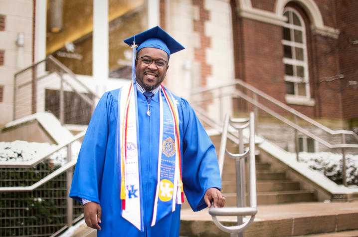 Graduate in a blue cap and gown with multiple honor cords and stoles, standing outside a brick building near steps on a snowy day.