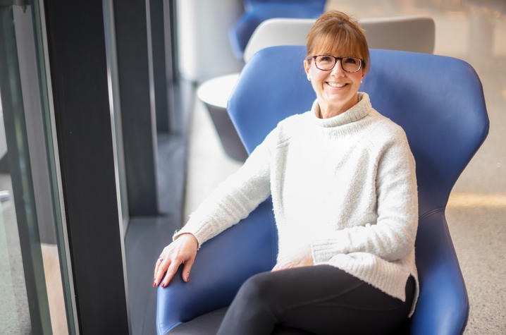 Photo of Jennifer Highland A woman wearing glasses and a light sweater sits smiling in a modern blue chair near a large window. Natural light fills the space, highlighting her relaxed posture in a bright, contemporary indoor setting.