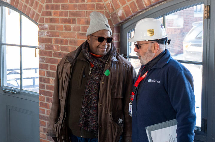 photo of Everertt McCorvey at Ellis Island Two people stand indoors near a brick wall and windows, talking. One wears a knit cap and jacket; the other wears a hard hat and holds a clipboard.