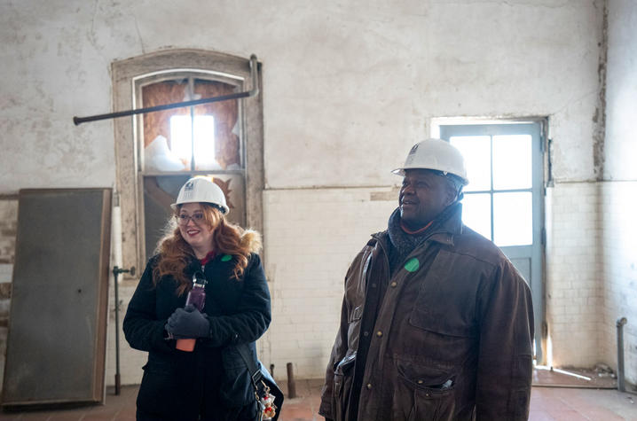 Megh Jennings-Posner and Everett McCorvey Two people wearing hard hats stand in a bright, unfinished room with brick and plaster walls.