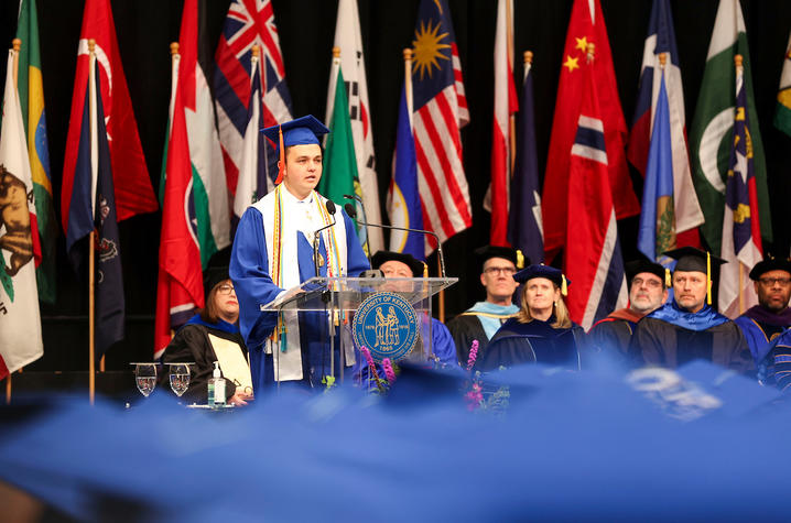 A student in a blue graduation cap and gown speaks at a podium on a stage, with faculty seated behind and numerous international flags displayed in the background.