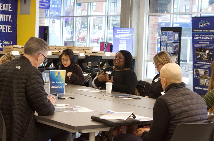Photo of participants at the UK Leadership Development Collaborative meeting. A group of people sit around a table in a bright meeting space, engaged in discussion. Papers, notebooks and a small sign stand on the table, and large windows and campus posters are visible in the background.