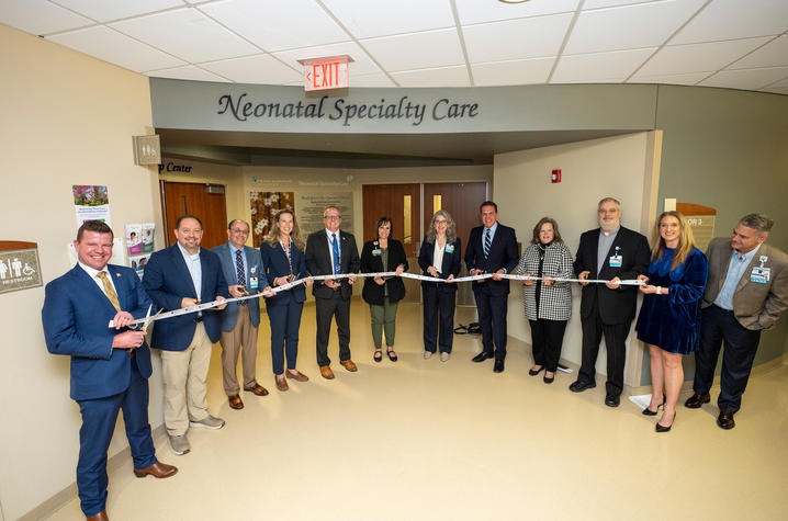 Photo of ribbon cutting at Saint Joseph London A group of people stand in a hallway under a sign reading ‘Neonatal Specialty Care,’ holding a long ribbon for a ribbon‑cutting ceremony.