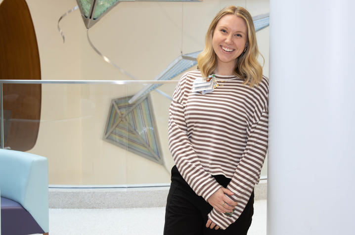 Image of Ella Kate Nichols in the lobby of UK Golisano Children’s A person wearing a striped long‑sleeve shirt and dark pants stands beside a white column in a modern indoor space with abstract artwork in the background