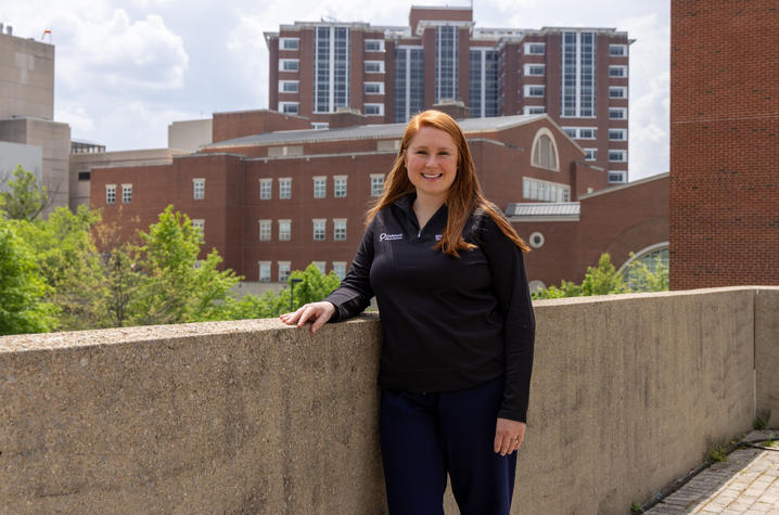 A person in black clothing stands outdoors with one hand resting on a concrete wall in front of red-brick campus buildings.