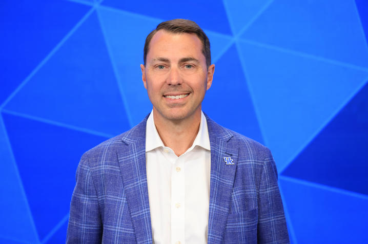 Smiling man in a blue jacket with a UK lapel pin against a blue geometric background.Provide your feedback on BizChat