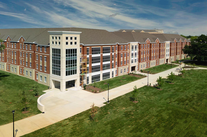 Aerial view of a large multistory brick academic building with a central tower, surrounded by sidewalks and open green lawn under a blue sky.