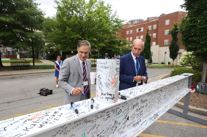 Two people stand outdoors beside a long steel beam covered with handwritten signatures and messages. Both wear business attire, with one in a light gray suit and patterned tie and the other in a blue suit and tie.
