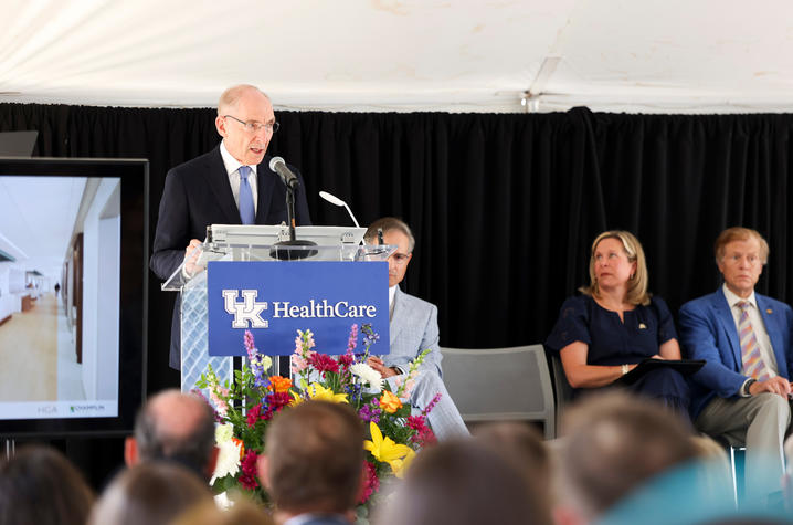A person speaks at a UK HealthCare podium decorated with flowers, with seated officials and an audience visible under a tent.