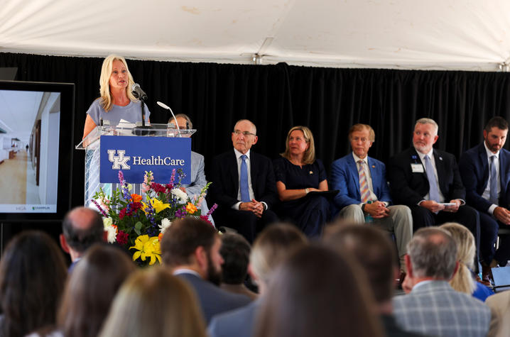 A person speaks at a UK HealthCare podium decorated with flowers, with seated officials and an audience visible under a tent.