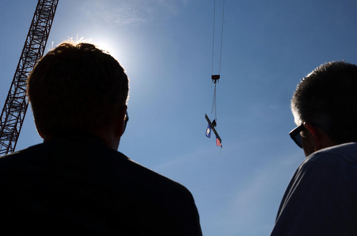 Two people in silhouette watch a crane lift a steel beam carrying an American flag and a Kentucky flag against a clear sky.