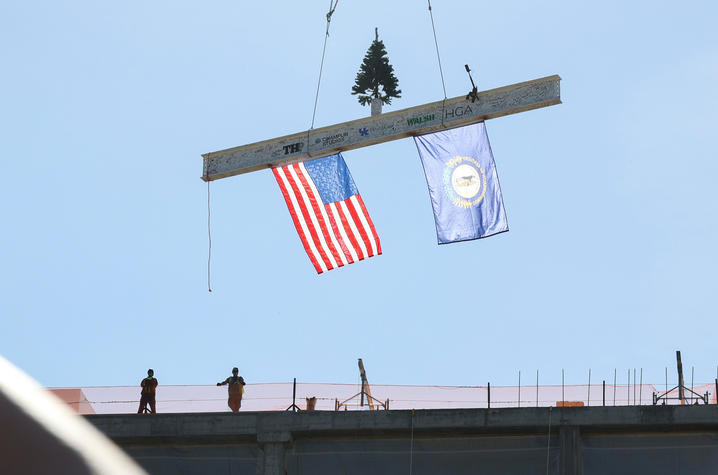 A steel beam signed by workers and decorated with a small tree, an American flag and a Kentucky flag is lifted by crane above a construction site, with two workers standing on the rooftop below.
