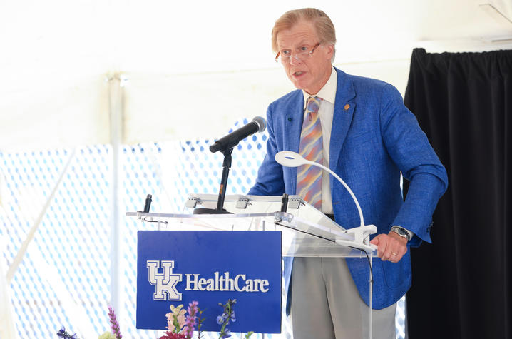 A person speaks at a UK HealthCare podium decorated with flowers, with seated officials and an audience visible under a tent.