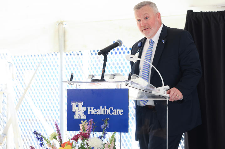 A person speaks at a UK HealthCare podium decorated with flowers, with seated officials and an audience visible under a tent.