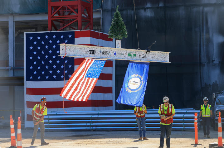 Workers guide a signed steel beam bearing an American flag and a Kentucky flag as a crane lowers it in front of a large flag mural at a construction site.