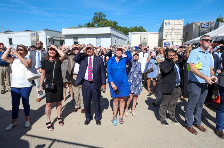 A crowd shields their eyes from the sun while looking up at a construction site, with some taking photos on their phones.