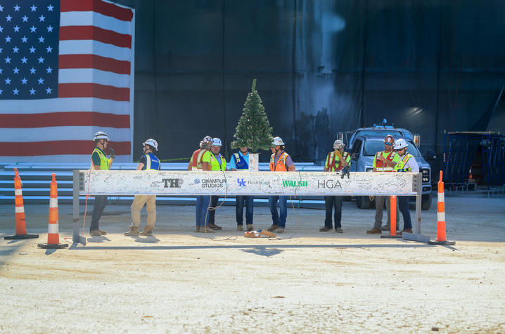 Construction workers in hard hats and safety vests stand around a signed steel beam topped with a small tree, with a large American flag mural in the background.