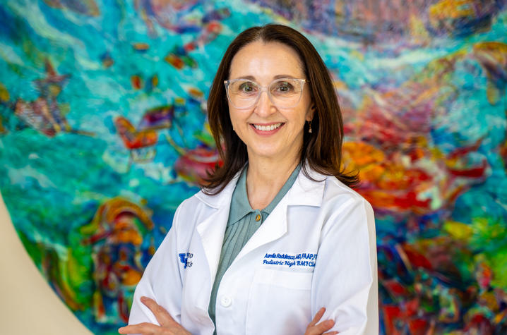 A health care provider in a white lab coat poses with arms crossed in front of a colorful abstract artwork. The portrait was taken in a clinical setting and highlights a leader in pediatric health care and research.