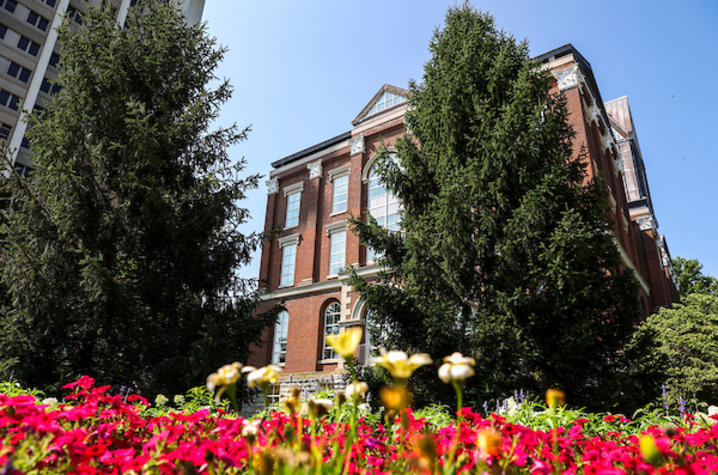 Historic brick building framed by flowers and evergreen trees.