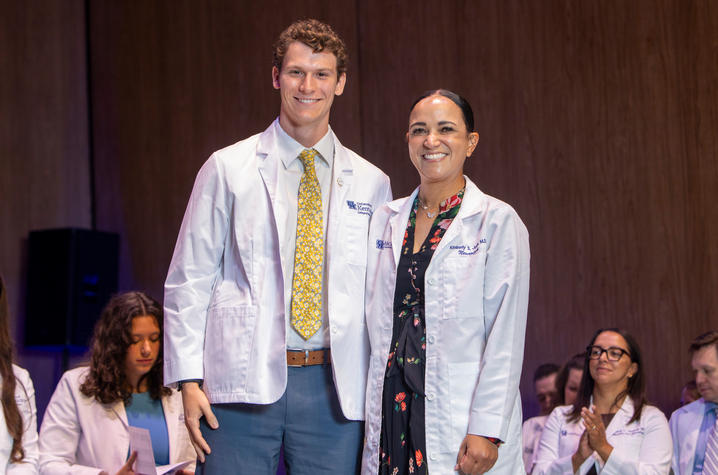 Two individuals wearing white coats stand together on a stage during a ceremonial event. One wears a light-colored shirt with a patterned yellow tie and blue trousers, while the other wears a dark patterned dress. 