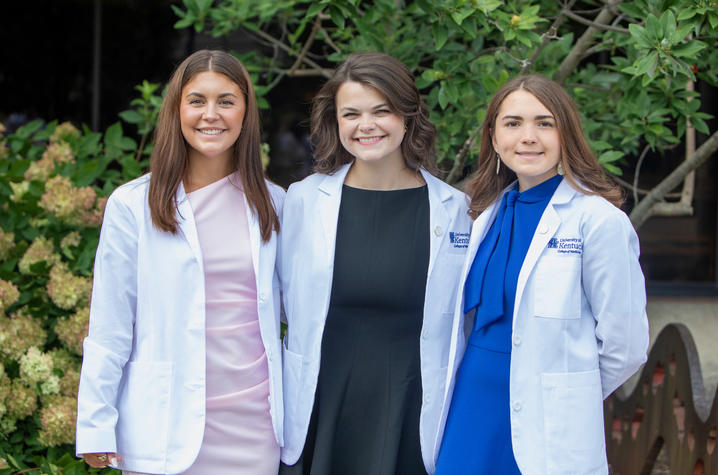 Three people wearing white coats stand side by side outdoors in front of green trees and flowering shrubs.