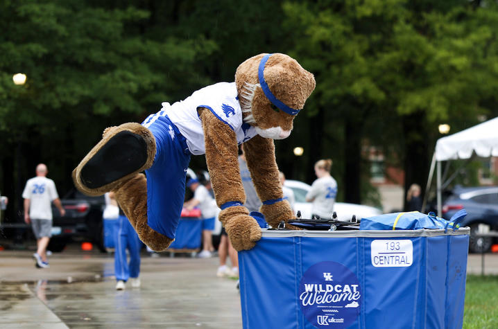 The Wildcat mascot playfully climbs onto a blue move-in cart on a rain-soaked campus sidewalk.