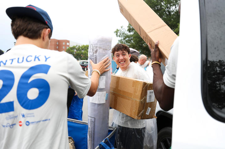 A smiling student in a clear rain poncho carries boxes as volunteers unload move-in supplies from a van.