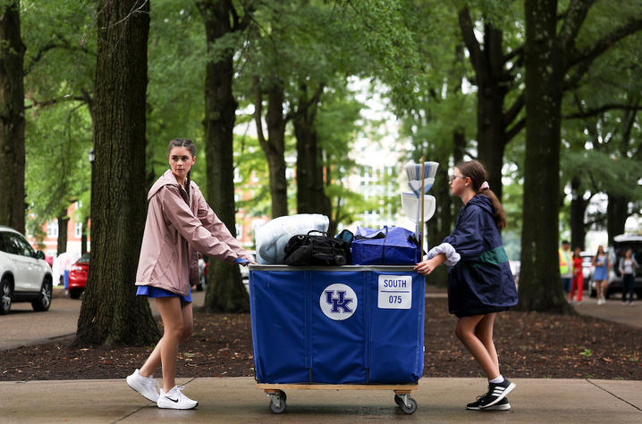 Two students push a blue cart loaded with dorm-room belongings along a tree-lined campus sidewalk during Big Blue Move-In.