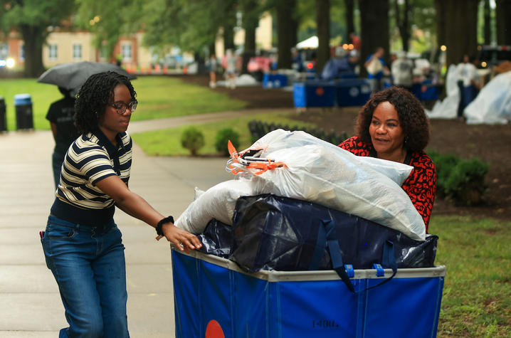 Two people push a blue cart piled with bags and bedding along a campus sidewalk during Big Blue Move-In.