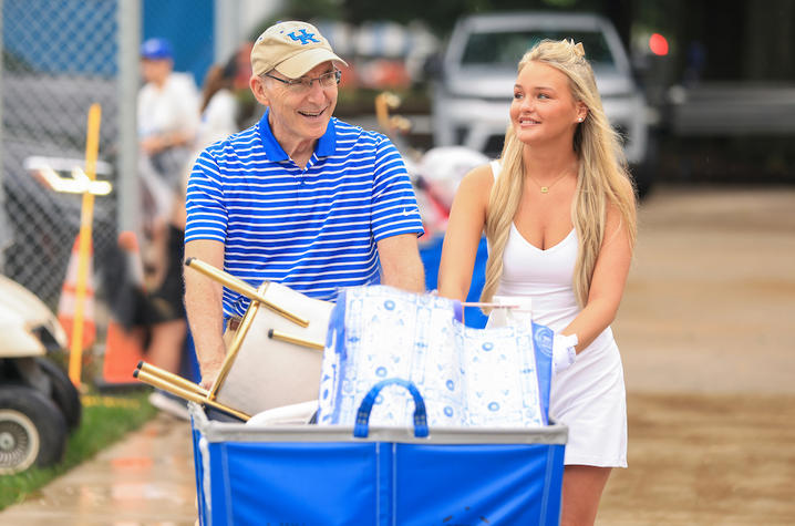 A student and man smile as they push a blue cart filled with dorm-room belongings during move-in.