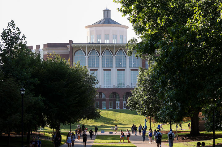 A large academic building stands at the end of a tree-lined campus walkway. Students walk along the path and gather on grassy areas beneath the trees on a sunny day.