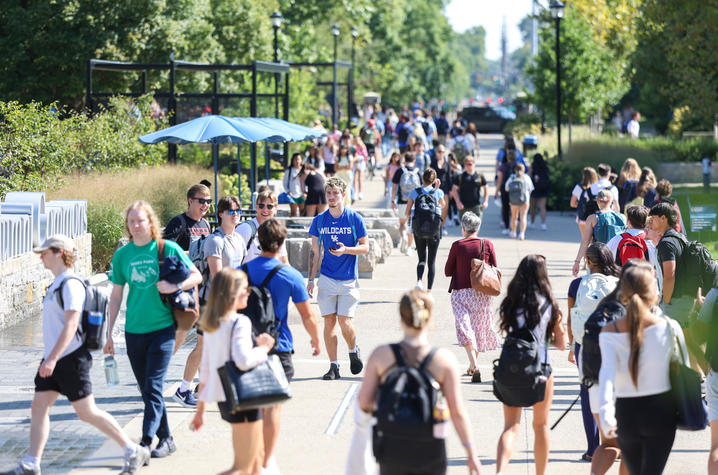 Students walk along a busy campus sidewalk on a sunny day during the start of the fall semester. People carrying backpacks travel in both directions along a tree-lined pedestrian pathway.
