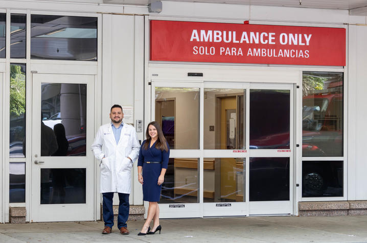 Two people stand outside a hospital ambulance entrance beneath a large red sign reading “AMBULANCE ONLY” and “SOLO PARA AMBULANCIAS.” One person is wearing a white medical coat, and the entrance doors and surrounding windows are visible in the background.