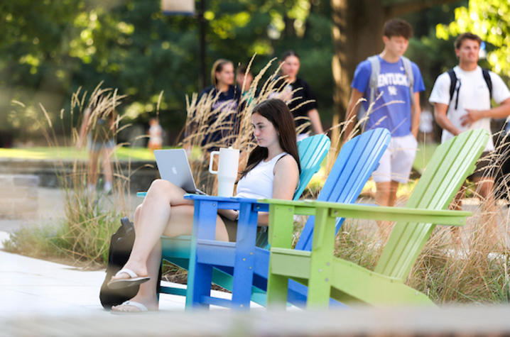  Person sitting in a colorful Adirondack chair outdoors, using a laptop and holding a mug while students walk nearby.