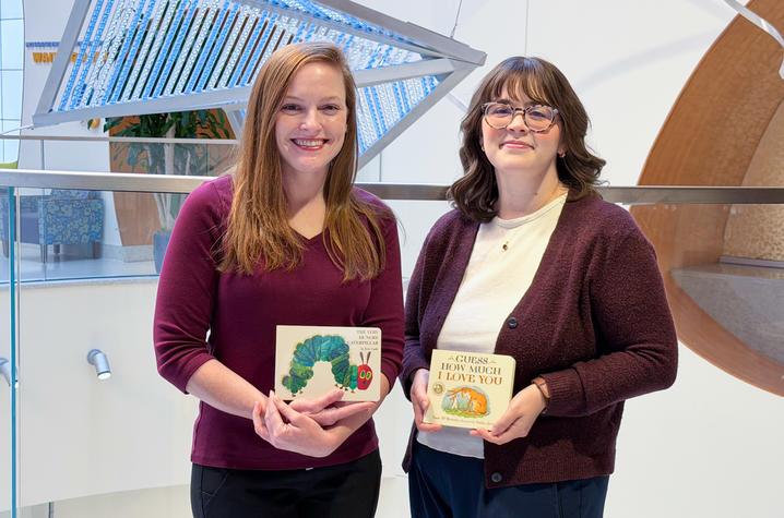 Two people stand indoors holding children’s books, with a large blue-lit art installation behind them.