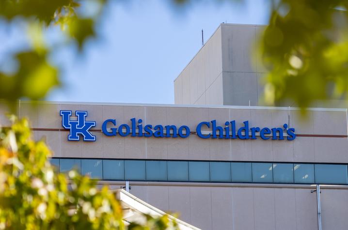 Exterior of the Golisano Children’s building featuring the blue UK logo and “Golisano Children’s” sign, framed by green tree leaves against a blue sky.