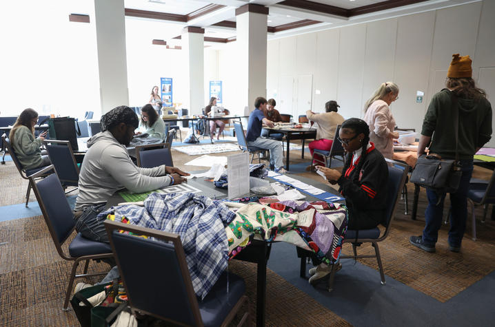 People sit at tables in a bright indoor room, working on sewing and fabric projects with machines and materials spread out.
