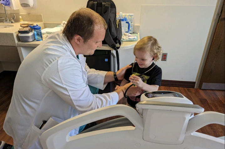 An adult wearing a white coat sits beside a hospital bed and holds a small medical instrument near a child seated in the bed, with medical supplies and a sink visible in the room.