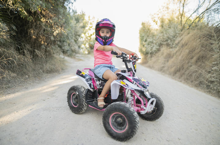 Child wearing a pink helmet and pink shirt sits on a small pink and black ATV on a dirt road lined with trees and dry grass.