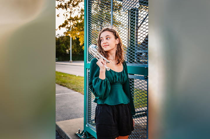 Woman in a green top outside, next to a metal structure, holding a trumpet. 